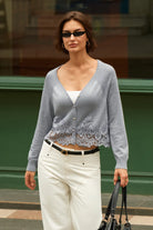 A woman wearing a light gray knit cardigan with a lace hem, white pants, and black sunglasses, holding a black handbag in an outdoor urban setting.