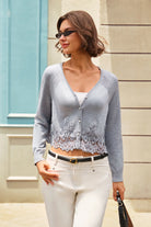 A woman wearing a light gray long-sleeve cardigan with a lace hem, styled with white pants and a black belt, standing outdoors against a beige building wall.
