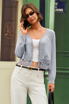 A woman models a light blue-gray cardigan with intricate lace applique at the hem, worn over a white crop top and white pants, standing in front of a green door.