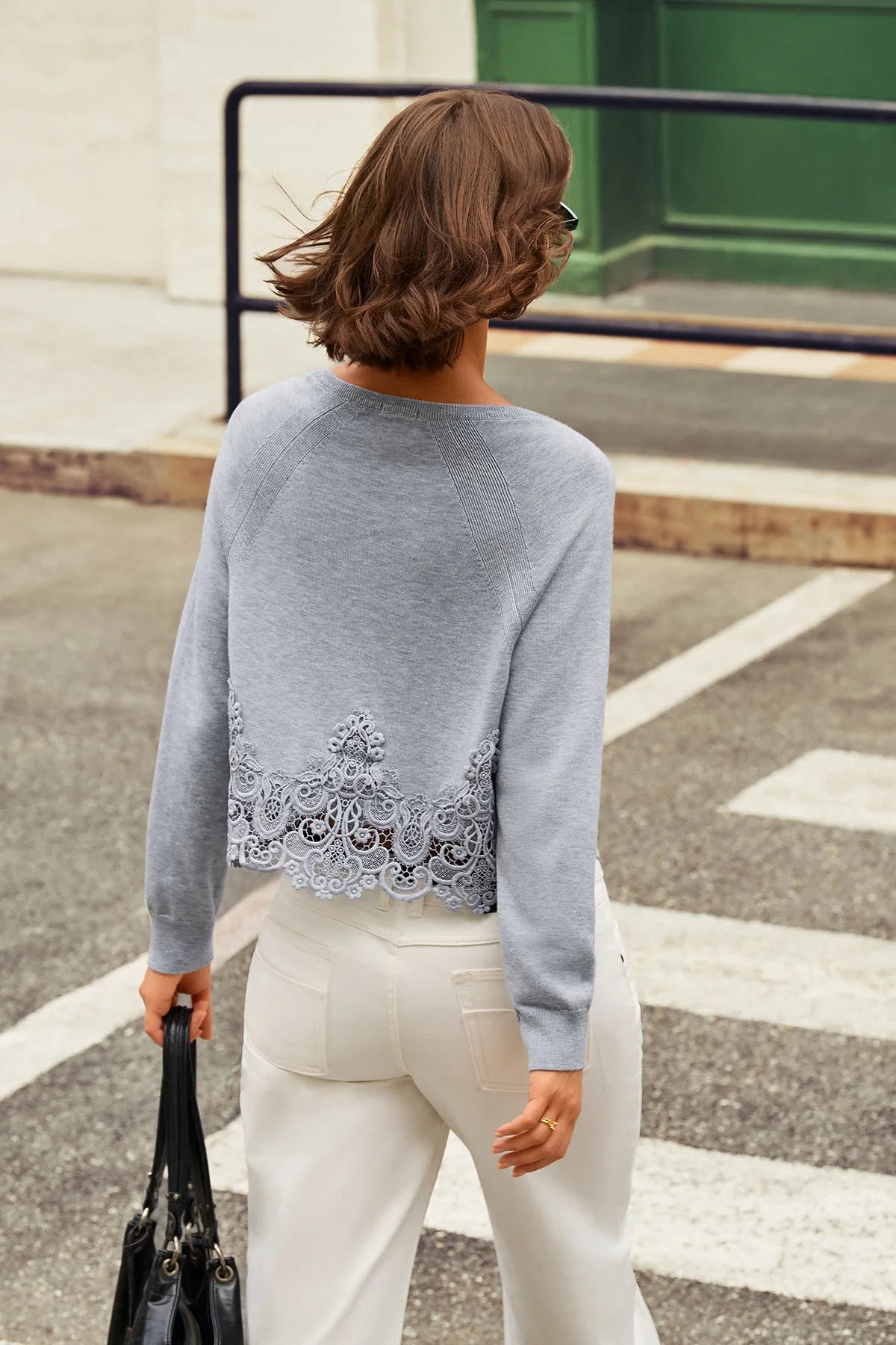 A woman viewed from behind wearing a light gray knit sweater with a decorative lace hem and white pants, standing on a street crosswalk.