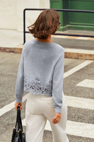 A woman viewed from behind wearing a light gray knit sweater with a decorative lace hem and white pants, standing on a street crosswalk.