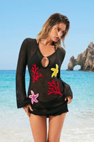 Woman wearing a black mesh swimsuit cover-up with colorful floral designs on a beach.