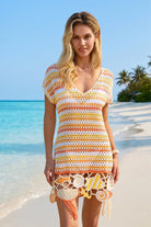 Woman in a colorful crochet dress standing on a beach with clear blue water and sky.