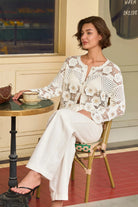 A woman sits on a woven chair at a small round table, wearing a white crochet cardigan and white wide-leg pants in an indoor cafe setting.