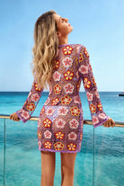 A woman wearing a long-sleeve mini dress with a colorful floral crochet lace pattern, standing on a balcony overlooking a tropical ocean under a clear blue sky.