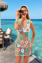A woman stands on a wooden deck by the ocean wearing a sleeveless crochet cover-up dress with a multicolor floral pattern. She is holding a camera up to her face, with turquoise water, a thatched roof, and outdoor seating visible in the background.