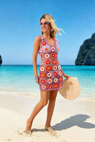 A woman stands on a sandy beach wearing a colorful crocheted mini dress with a floral pattern, holding a wide-brimmed straw hat. The background features turquoise ocean water, a blue sky, and a green rocky island.