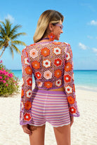 Woman in a colorful crochet outfit on a beach with palm trees and blue sky.
