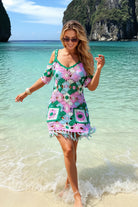 A woman wearing a colorful crochet mini dress walks into shallow turquoise ocean water on a sandy beach, with tropical cliffs in the background.