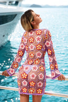 Woman in a colorful floral dress standing by a body of water with a boat in the background.