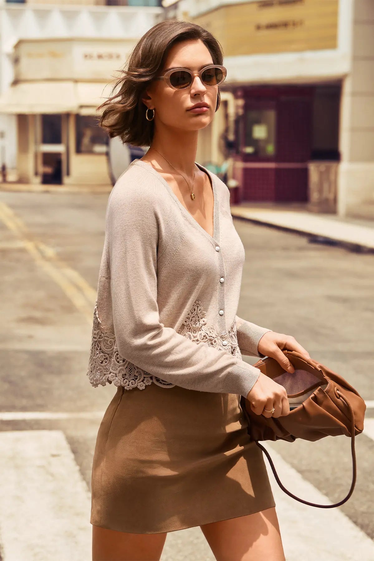 A woman walking on a city street wearing a light beige knit cardigan with lace trim and a brown mini skirt, holding a brown leather handbag.