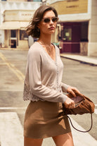 A woman walking on a city street wearing a light beige knit cardigan with lace trim and a brown mini skirt, holding a brown leather handbag.