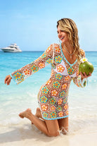 A woman kneeling in shallow ocean water on a beach wears a long-sleeve crochet cover-up dress with a colorful floral pattern over a white bikini. She holds a green coconut, with a blue sky and a white yacht in the background.