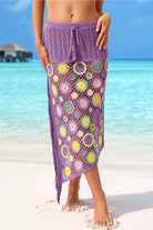 Woman standing on sandy beach wearing twilight purple crochet maxi skirt with colorful 3D floral motifs and drawstring tassel waist, turquoise ocean in background